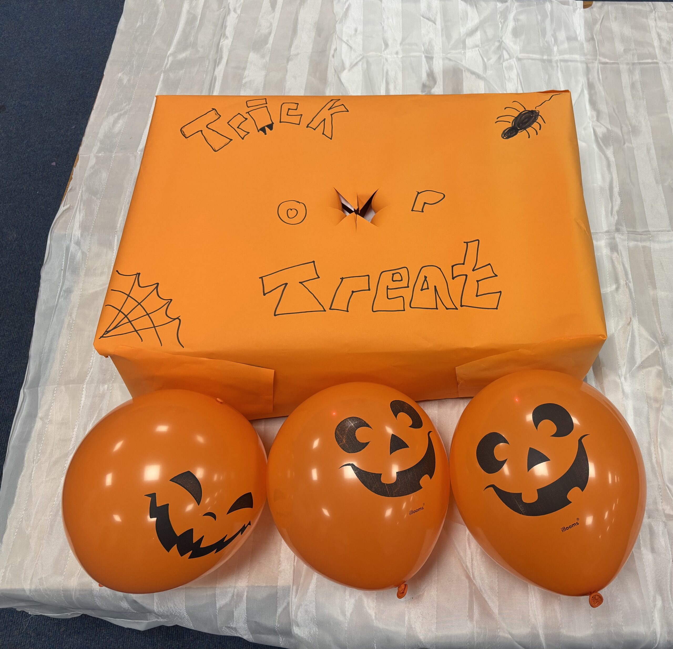 Harlow Halloween Bright orange “Trick or Treat” box decorated with Halloween drawings, surrounded by pumpkin-face balloons, created by children at Action for Family Carers’ Harlow Young Carer Club Halloween session.